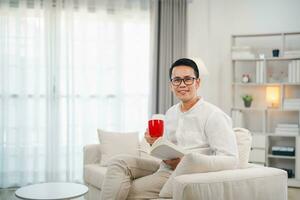 Joyful asian man reading book and drink hot coffee while sitting on couch sofa in living room at home. Asian man relex reading book smile at sofa In the house. Activity hobby at house concept. photo