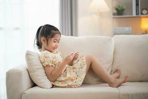 Asian child girl looking using and touch mobile phone screen on couch sofa. Baby smiling funny time to use mobile phone. Too much screen time. Cute girl watching videos while tv, Internet addiction. photo