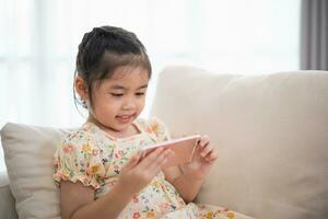 Asian child girl looking using and touch mobile phone screen on couch sofa. Baby smiling funny time to use mobile phone. Too much screen time. Cute girl watching videos while tv, Internet addiction. photo