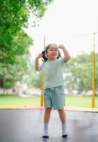 Asian child girl is jumping on trampoline on playground background. Happy laughing kid outdoors in the yard on summer vacation. Jump high on trampoline. Activity children in the kindergarten school. photo