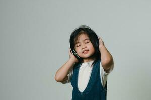 Portrait of relaxed cute little asian girl child wearing white headphones, keeps hands on her ears, posing and smiling enjoying isolated over gray background. photo