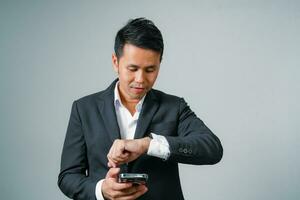 Asian businessman in suit with using mobile and looking at watch, standing studio isolated on gray background, young business man is manager or executive smiling and confident is positive success. photo