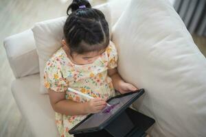 Asian child girl using pen and touch drawing on tablet display screen. Baby smiling funny time to use tablet. Too much screen time. Cute girl watching videos while tv, Internet addiction concept. photo