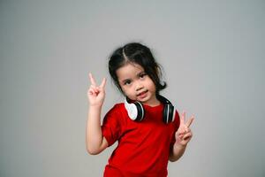 alegre contento asiático niños niña con negro pelo escuchando a música canciones en auriculares en un gris antecedentes. retrato linda niña advirtiendo rojo camisa y mirando a cámara estudio Encendiendo. foto