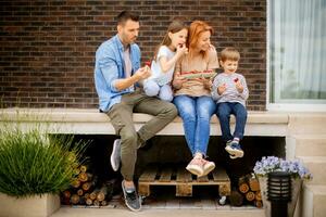 familia con un madre, padre, hijo y hija sentado fuera de en pasos de un frente porche de un ladrillo casa y comiendo fresas foto