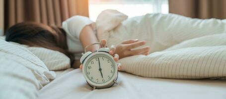 alarm clock and asian woman hand stop time in bed while sleeping, young adult female wake up late in the early morning. Relaxing , sleepy, daily routine and have a nice day concepts photo
