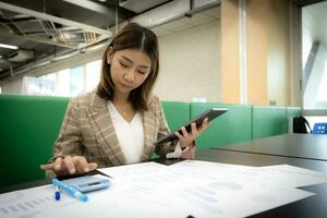 Beautiful asian businesswoman sitting and focusing on documents in the company while one hand is holding a tablet and the other is pressing a calculator. photo