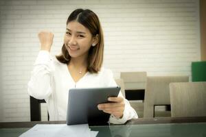 Seated in a close-up shot the beautiful asian businesswoman wearing white dress is happily displaying a fist while holding a tablet  pleased with the sales from her online store. photo