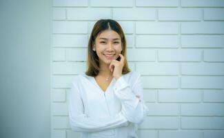 Beautiful asian business woman is standing with her hand on her cheek and smiling with a white brick wall in the background, Digital marketing. photo