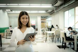 Beautiful asian female teacher is standing and looking to the camera while holding tablet in the library room. photo