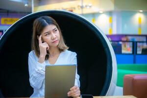 hermosa asiático negocio mujer es sentado en un moderno redondo silla con un tableta en su mano y un interrogatorio Mira en su rostro, digital marketing. foto