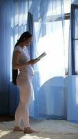 un' incinta donna nel bianca pantaloni e un' bianca camicia è in piedi nel davanti di un' finestra lettura un' libro video