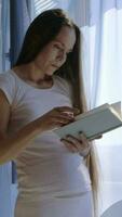 un embarazada mujer en blanco pantalones y un blanco camisa es en pie en frente de un ventana leyendo un libro video