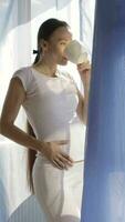 a pregnant woman in white pants drinking coffee while standing by the window video