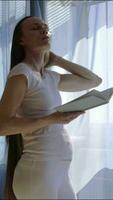 un embarazada mujer en blanco pantalones y un blanco camisa es en pie en frente de un ventana leyendo un libro video