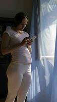 a pregnant woman in white pants and a white shirt standing by the window is using her phone video