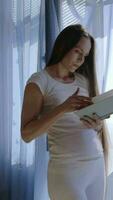 un embarazada mujer en blanco pantalones y un blanco camisa es en pie en frente de un ventana leyendo un libro video