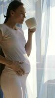 een zwanger vrouw in wit broek drinken koffie terwijl staand door de venster video