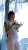 uma grávida mulher dentro branco calça e uma branco camisa é em pé dentro frente do uma janela lendo uma livro video