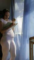 a pregnant woman in white pants and a white shirt is standing in front of a window reading a book video