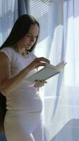 een zwanger vrouw in wit broek en een wit overhemd is staand in voorkant van een venster lezing een boek video