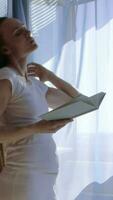 un embarazada mujer en blanco pantalones y un blanco camisa es en pie en frente de un ventana leyendo un libro video