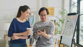 dos joven asiático puesta en marcha empresa socios discutir y idea genial en casual pequeño negocio oficina, márketing planificación trabajos en un tableta, y Lanza libre trabajo como comercio electrónico empresario y compañero de trabajo equipo. video