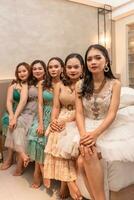 a group of Asian women sitting together on a white bed while wearing dresses and makeup during a party at a friend's house photo