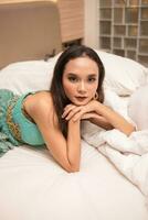 un hermosa asiático mujer es relajante en un blanco cama mientras vistiendo maquillaje y vestir foto