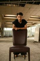 an Asian man posing on a brown leather chair while wearing black pants in an old building photo