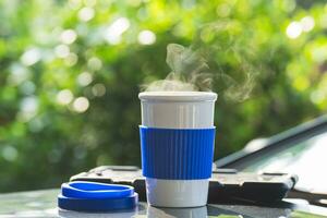 Hot ceramic white coffee cup with smoke placed outside the car dashboard at sunrise in the morning, in a natural background. Hot Coffee Drink Concept, selective focus, soft focus. photo