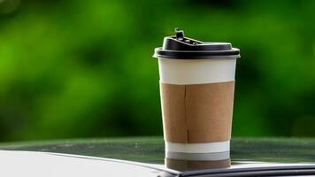 café para llevar en un papel taza en parte superior de el coche techo verde árbol antecedentes a amanecer en el mañana, selectivo enfocar, suave enfocar. foto