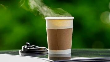 café para llevar en un papel taza en parte superior de el coche techo verde árbol antecedentes a amanecer en el mañana, selectivo enfocar, suave enfocar. foto