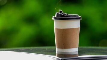 coffee takeaway in a paper cup on top of the car roof green tree background at sunrise in the morning,  selective focus, soft focus. photo