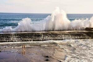 two people standing on the beach near a large wave photo