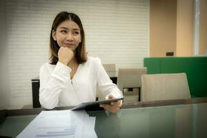 Beautiful asian office girl is sitting pretending to think in copy space with a doubtful expression and holding tablet on her desk. photo