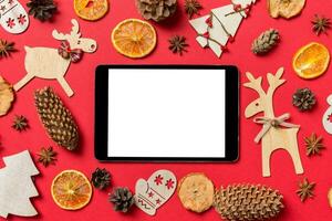 Top view of digital tablet, red background decorated with festive toys and Christmas symbols reindeers and New Year trees. Holiday concept photo