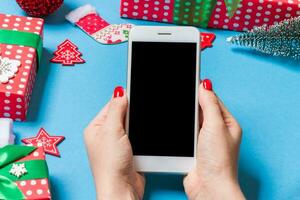 Top view of phone in female hand on festive blue background. Christmas decorations. New Year time holiday. Mockup photo