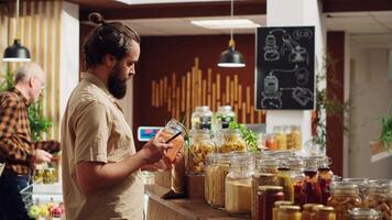 Zero waste supermarket customer shopping for pantry staples, using smartphone to take photo for wife at home. Man in local neighborhood shop using mobile phone to agree on what to buy with partner video