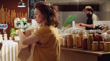 Woman adding organic pantry staples and vegetables in paper bag, getting product recommendations from vendor in zero waste supermarket. Seller helping client in local neighborhood shop video