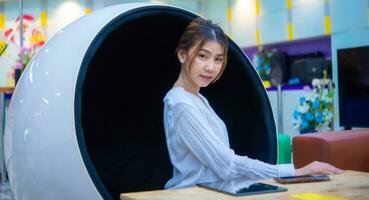 Beautiful asian business woman sitting on a modern round chair touching the table and has a phone and tablet on it and looking at the camera, Digital marketing. photo