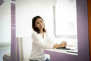 Beautiful asian businesswoman coming to work happy while looking at camera and sitting in her workplace. photo