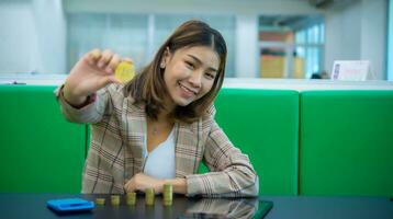 hermosa asiático negocio mujer es sentado con uno mano participación un oro moneda elevado en frente de mientras allí un pila de monedas un calculadora y un tableta en negro mesa, digital marketing. foto