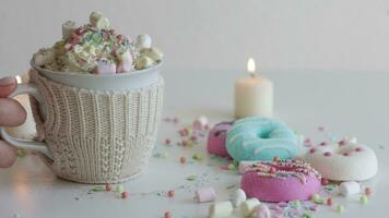 Nahansicht Frau Hand setzt ein Becher auf das Tisch. Tasse von heiß Schokolade mit Marshmallows. bunt Donuts auf ein Weiß Tabelle umgeben mit Kerzen video