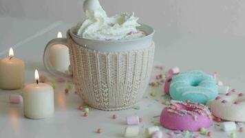 Putting whipping cream into glass mug with a hot chocolate and marshmallows. Colorful donuts on a white table surrounded with candles at home video