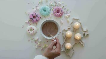 Top view of cocoa being stirred with a golden whisk with colorful marshmallows. Colorful donuts on a white table surrounded with candles video