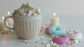 Close-up woman's hand puts a mug on the table. Cup of hot chocolate with marshmallows. Colorful donuts on a white table surrounded with candles video