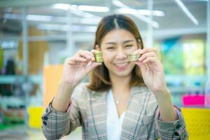 hermosa asiático negocio mujer radiante sonrisa brilló como ella sostener un reluciente oro moneda en su mano. foto