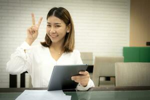 Beautiful asian wearing white dress in close-up shot is sitting showing victory sign while holding tablet with smile on her face, Business concept. photo