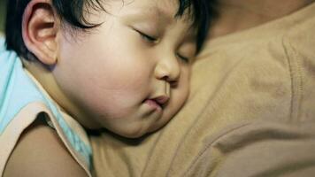 pequeño chico dormido en su papá. padre participación su pequeño hijo en su brazos sentado en el sofá en el casa. familia concepto. video